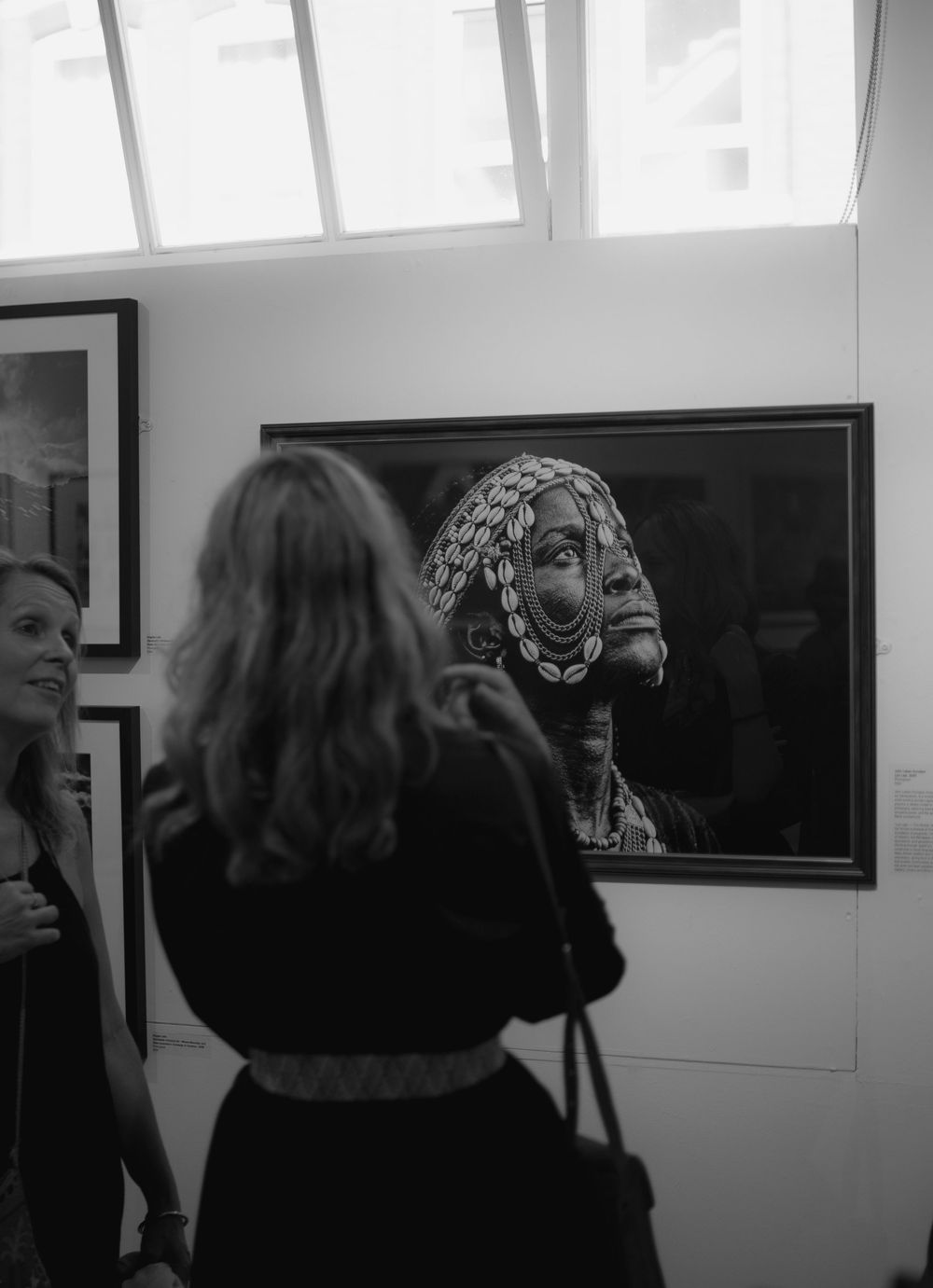 Two women admire a framed black and white portrait of a decorated tribal figure at an art gallery.