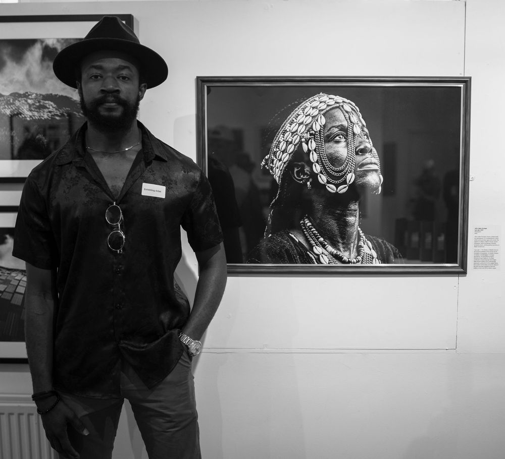 Black and white photo of a man in a hat standing beside a framed portrait photograph in an art gallery.