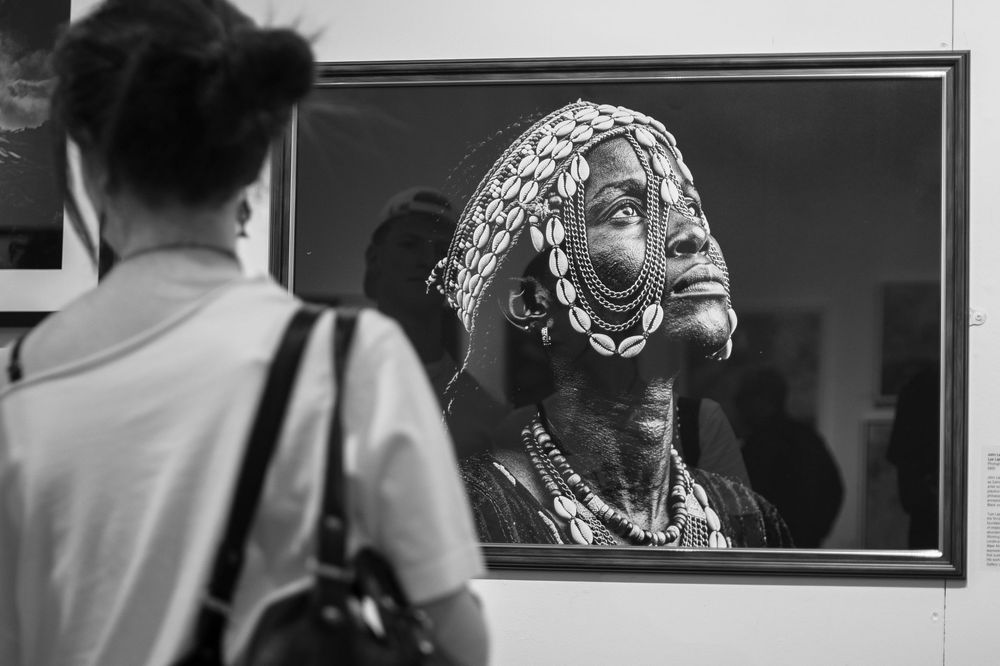 Black and white photo of a person viewing a framed portrait of a traditionally adorned African woman at an art gallery.