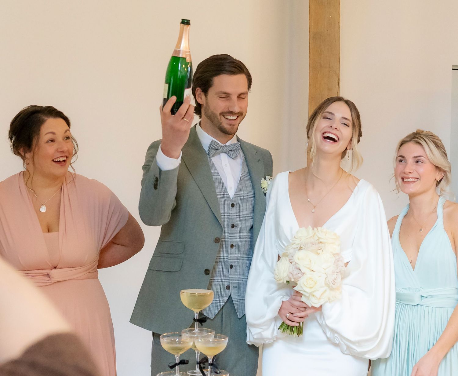 Joyful bride and groom celebrating with champagne bottle alongside smiling bridesmaids at wedding reception.