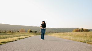 Woman in black hat and jeans standing alone on rural road with mountain backdrop.