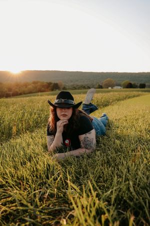 A tattooed woman in a black hat lies in a lush green field during golden hour.