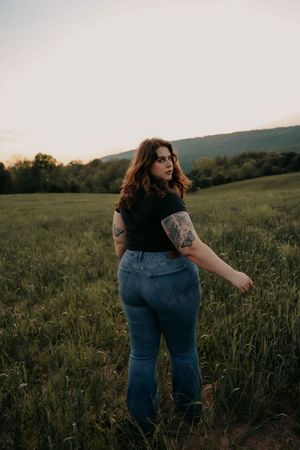 Woman with tattoos in black top and jeans poses in a green meadow during a beautiful golden hour sunset.