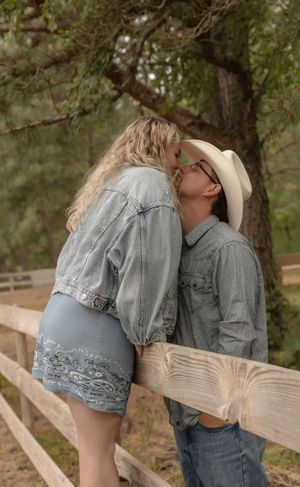 Couple sharing a romantic kiss leaning against a wooden fence with lush green trees in the background.