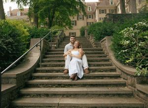 Couple sitting together on stone outdoor staircase surrounded by lush greenery and historic building backdrop.
