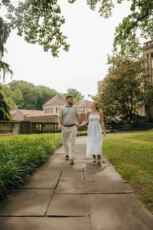 A couple holds hands walking on a stone path through a lush green campus garden with historic buildings.