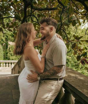 A couple embraces romantically under lush green trees on a stone balcony, the man tattooed, both dressed in neutral tones.