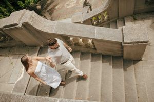 Couple in white outfits holding hands while walking up elegant stone stairs, viewed from above.