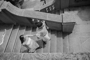 Black and white aerial view of a couple holding hands while walking up stone stairs together.