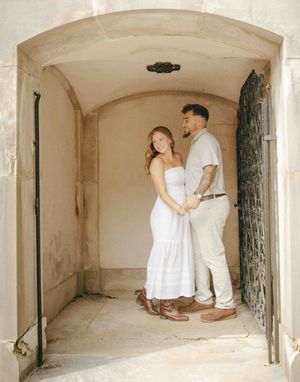 Couple in white outfits holding hands under stone archway with ornate iron door for engagement photo.