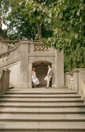 Couple in white attire standing on stone steps beneath lush trees at an elegant outdoor venue.