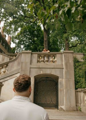 A man looks up at a woman leaning on a stone balustrade of an elegant mansion staircase surrounded by trees.