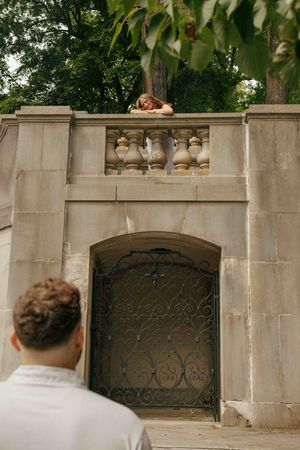 A woman leans over a stone balcony above an ornate iron gate, looking down at a man in a white shirt below.