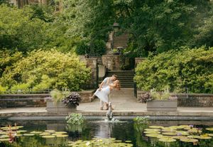 Couple sharing a romantic dip kiss by a lily pond in a lush garden with stone steps and fountain.
