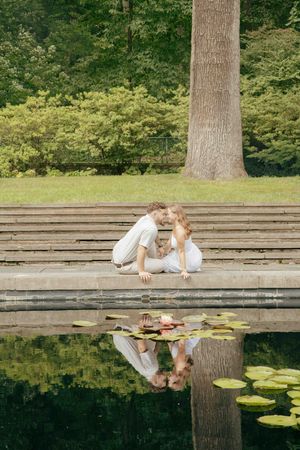 Couple sharing a romantic moment by a lily pad pond on stone steps in a lush green park.