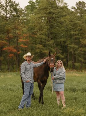Couple in denim standing with brown horse in green meadow surrounded by colorful autumn trees.