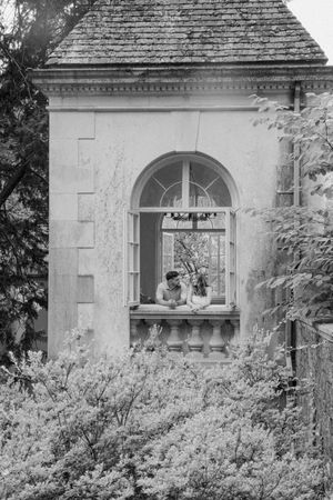 Black and white photo of a couple leaning on a balustrade in an arched window of a historic stone building.