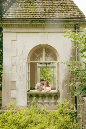 Couple sharing a romantic moment in an arched window of a vintage stone building surrounded by lush greenery.