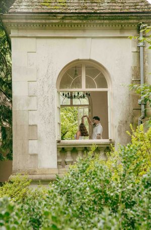 Couple standing on a balcony of an elegant stone building surrounded by lush green gardens.