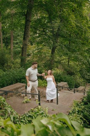 Couple in white outfits holding hands on stone steps surrounded by lush green trees in a romantic garden setting.