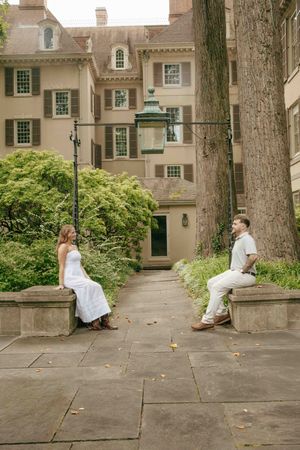 A couple in white attire sits on stone benches facing each other in an elegant courtyard with lush greenery.