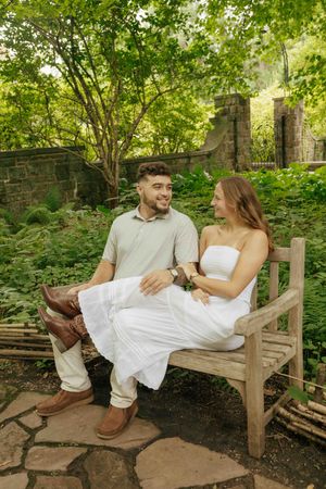 A smiling couple sits on a wooden bench in a lush green garden, the woman in white dress, man in beige outfit.