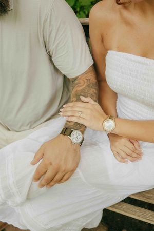 Couple sitting on bench showing matching watches, tattoos, engagement ring, and coordinating white outfits.