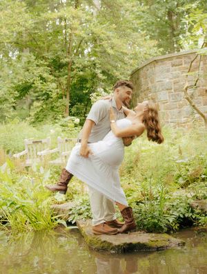Couple sharing a romantic dip pose on stepping stones in a lush garden, woman in white dress, man in casual attire.