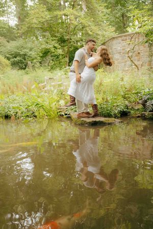 Couple sharing a romantic moment by a koi pond in a lush garden, with their reflection visible in the water.