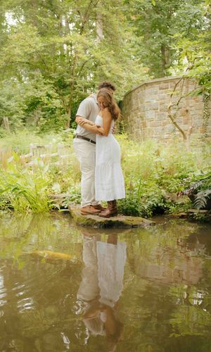 A couple embraces romantically by a tranquil garden pond, their reflection visible in the water, surrounded by lush greenery.