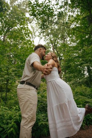 A couple in white and beige outfits dancing romantically in a lush green forest setting.