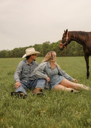 A couple sits in a grassy field with a brown horse nearby on a cloudy day.