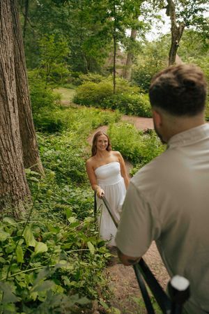 Bride in white strapless dress smiles at groom on a lush green forest path during an outdoor wedding photo session.