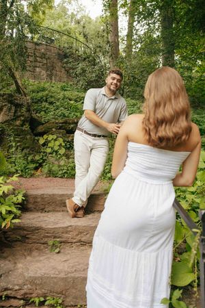 A smiling man walks up stone steps toward a woman in a white dress in a lush green garden setting.
