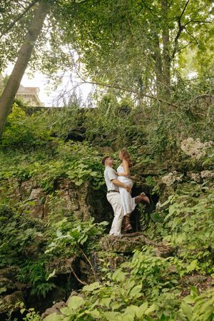A couple in white wedding attire embraces romantically on a rocky ledge surrounded by lush green forest foliage.