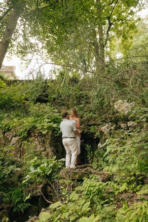 Couple embracing on mossy rocks surrounded by lush green trees and foliage in a romantic outdoor setting.
