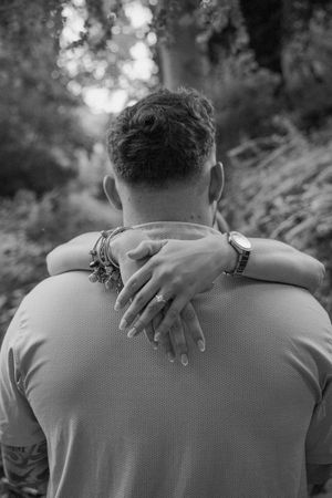 Black and white photo of a couple embracing outdoors, woman's hands with ring and bracelets around man's neck.