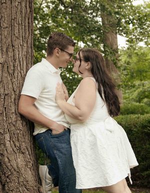 A couple shares a romantic moment leaning against a tree in a lush green park, dressed in white and jeans.