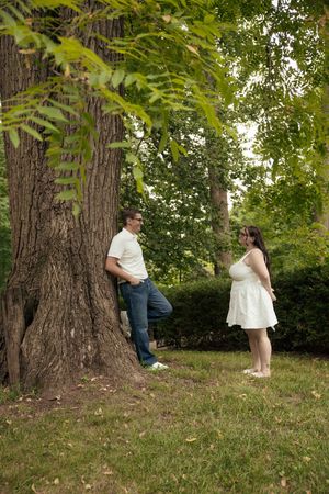 Pregnant woman in white dress and man in white shirt chat near large tree in lush green park.