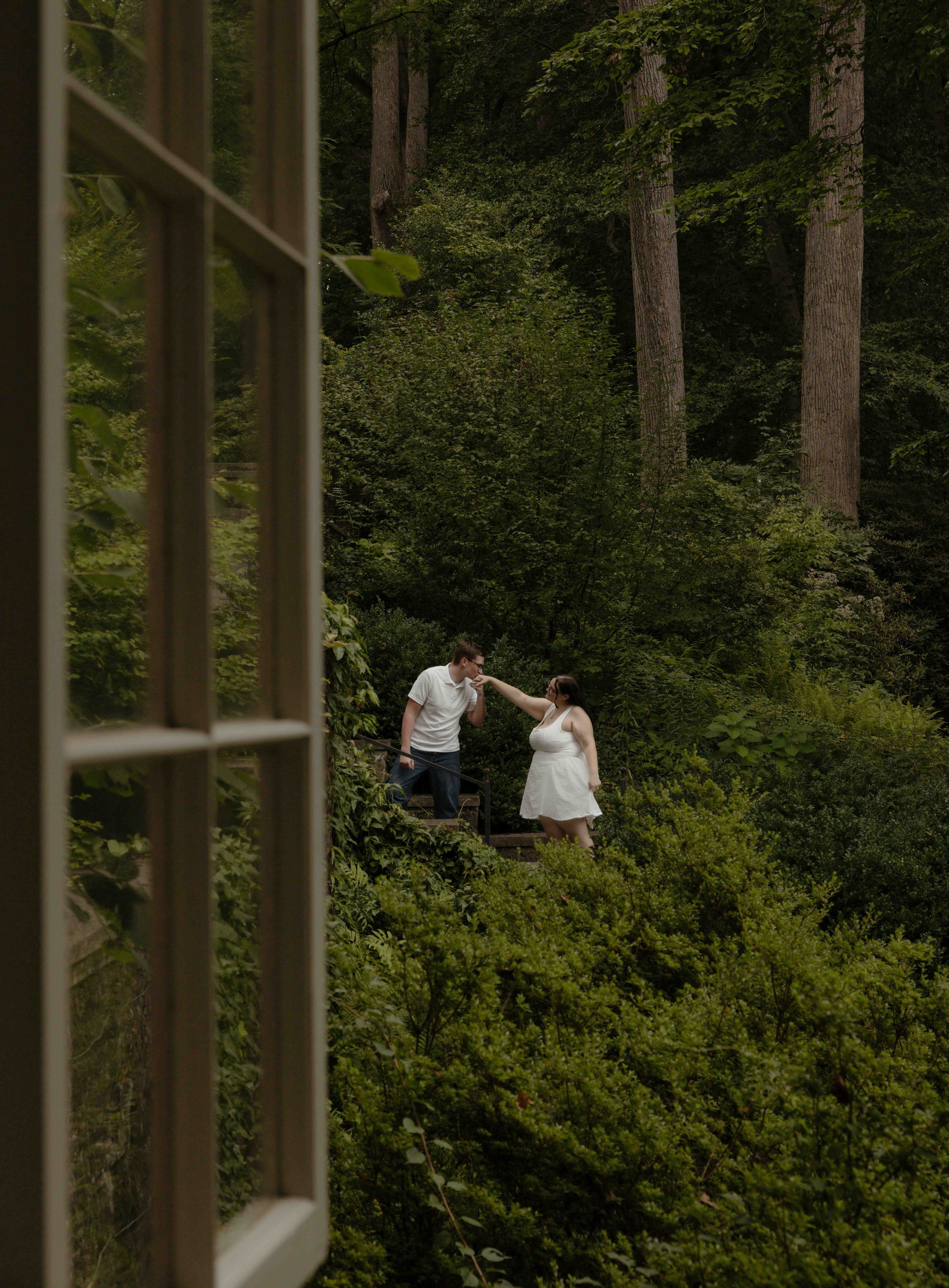 Couple dances in lush green forest viewed through open white window frame.