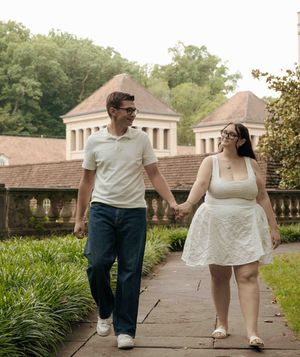 Couple holding hands walking on a brick path in a lush garden with a historic building backdrop.