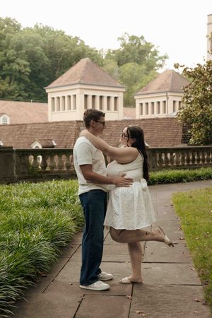 A couple embraces on a garden path, the woman in a white dress lifting one leg, with a historic building behind them.