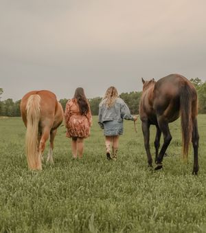 Two women walking with horses through green field under cloudy sky at dusk.