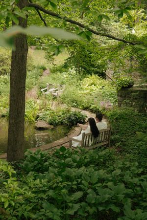 Couple sitting on a wooden bench in a lush green garden beside a tranquil pond surrounded by trees.