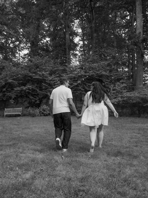 Black and white photo of a couple holding hands walking through a lush green park toward tall trees.