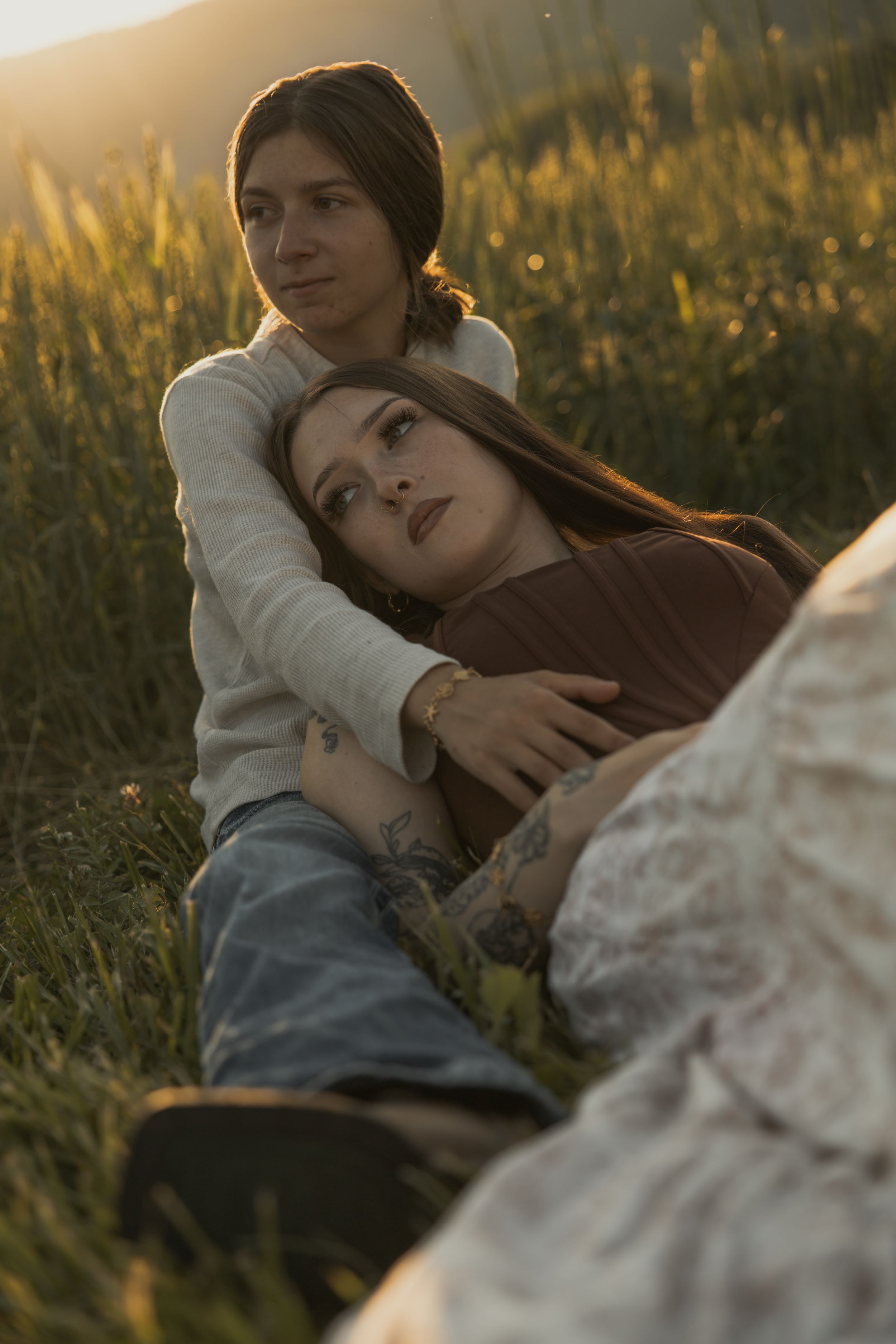 Two women relaxing in golden sunlit meadow, one resting her head on the others lap.