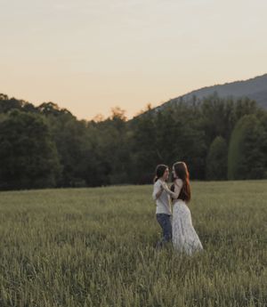 Two women embrace in a green field with mountains and trees at sunset.