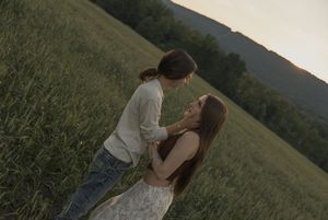 Two young women share a tender moment in a lush green field at sunset with mountains in the background.