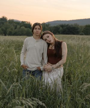 Young couple standing together in a tall grass field at sunset with rolling hills in the background.
