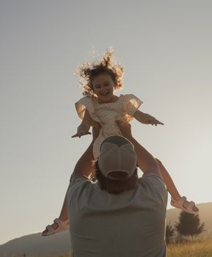 Joyful father lifting smiling little girl in floral dress against golden sunset sky.